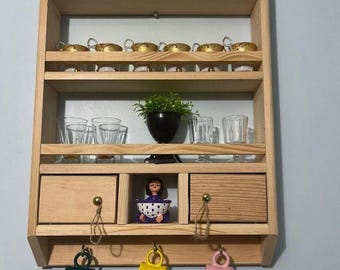 May include: A wooden wall-mounted shelf with three shelves, two drawers, and hooks for hanging mugs. The shelves are filled with various cups and glasses. There is a small doll figurine in the center drawer.