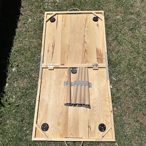 May include: A rectangular wooden cornhole board with a natural wood finish. The board has black corner protectors and a metal handle. The legs are folded and secured with a gray strap. The board is set on green grass.