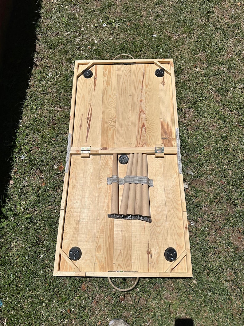 May include: A rectangular wooden cornhole board with a natural wood finish. The board has black corner protectors and a metal handle. The legs are folded and secured with a gray strap. The board is set on green grass.