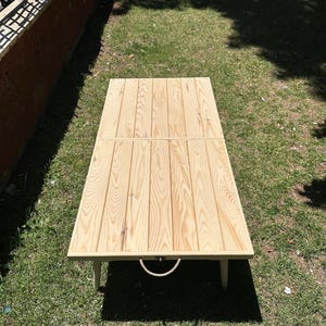 May include: A rectangular wooden table with a light brown finish. The table is made of vertical wooden planks and has a rope handle. The table is on a grassy surface.