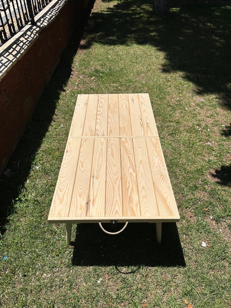 May include: A rectangular wooden table with a light brown finish. The table is made of vertical wooden planks and has a rope handle. The table is on a grassy surface.
