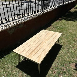May include: A rectangular wooden table with a light-colored, natural wood finish. The table has four legs and a rope handle. The table is approximately 48 inches long and 24 inches wide. The table is sitting on green grass.