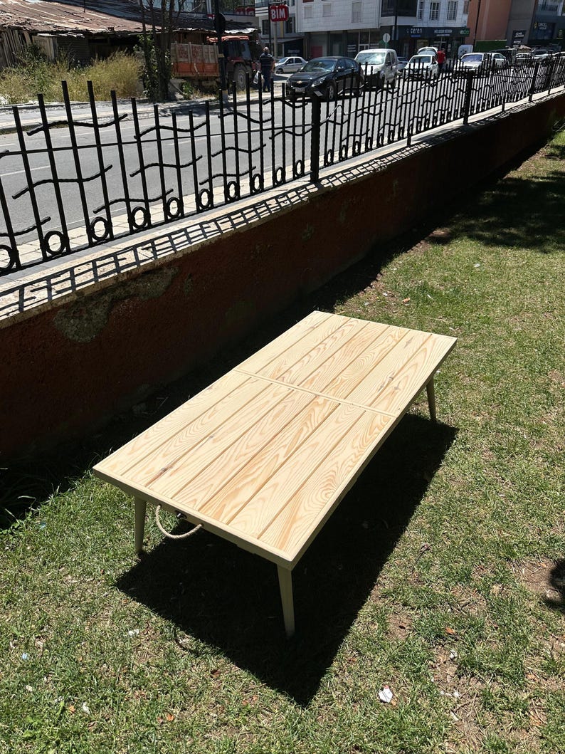 May include: A rectangular wooden table with a light-colored, natural wood finish. The table has four legs and a rope handle. The table is approximately 48 inches long and 24 inches wide. The table is sitting on green grass.