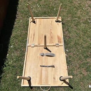 May include: A light-colored wooden cornhole board with four wooden legs and a handle. The board is rectangular with a hole in the top surface. The legs are attached with black rubber feet. The board is set on a grassy surface.