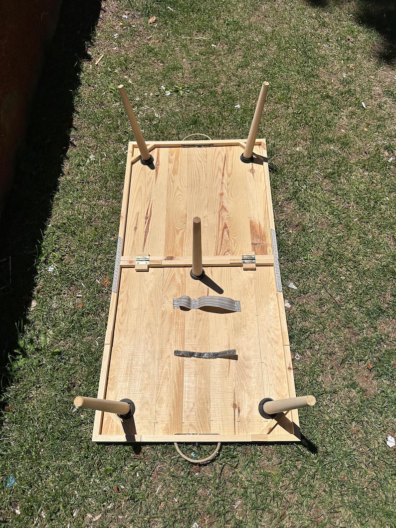 May include: A light-colored wooden cornhole board with four wooden legs and a handle. The board is rectangular with a hole in the top surface. The legs are attached with black rubber feet. The board is set on a grassy surface.