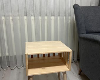 Puede incluir: Una mesa auxiliar de madera clara con un estante y cuatro patas. La mesa está en una habitación con cortinas blancas y un sillón gris.