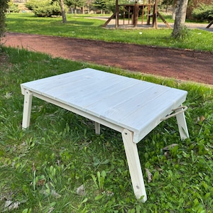 Può includere: Un tavolo pieghevole in legno bianco con un design semplice. Il tavolo è installato su un prato.