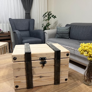 Op de afbeelding: Een houten kist met een scharnierend deksel en zwarte metalen accenten. De kist is gemaakt van licht hout en heeft een rustieke uitstraling.