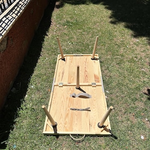 May include: A rectangular wooden picnic table with four detachable legs. The table is made of light-colored wood and has a rope handle. A fork and knife are placed on the table. The table is set on a grassy area.