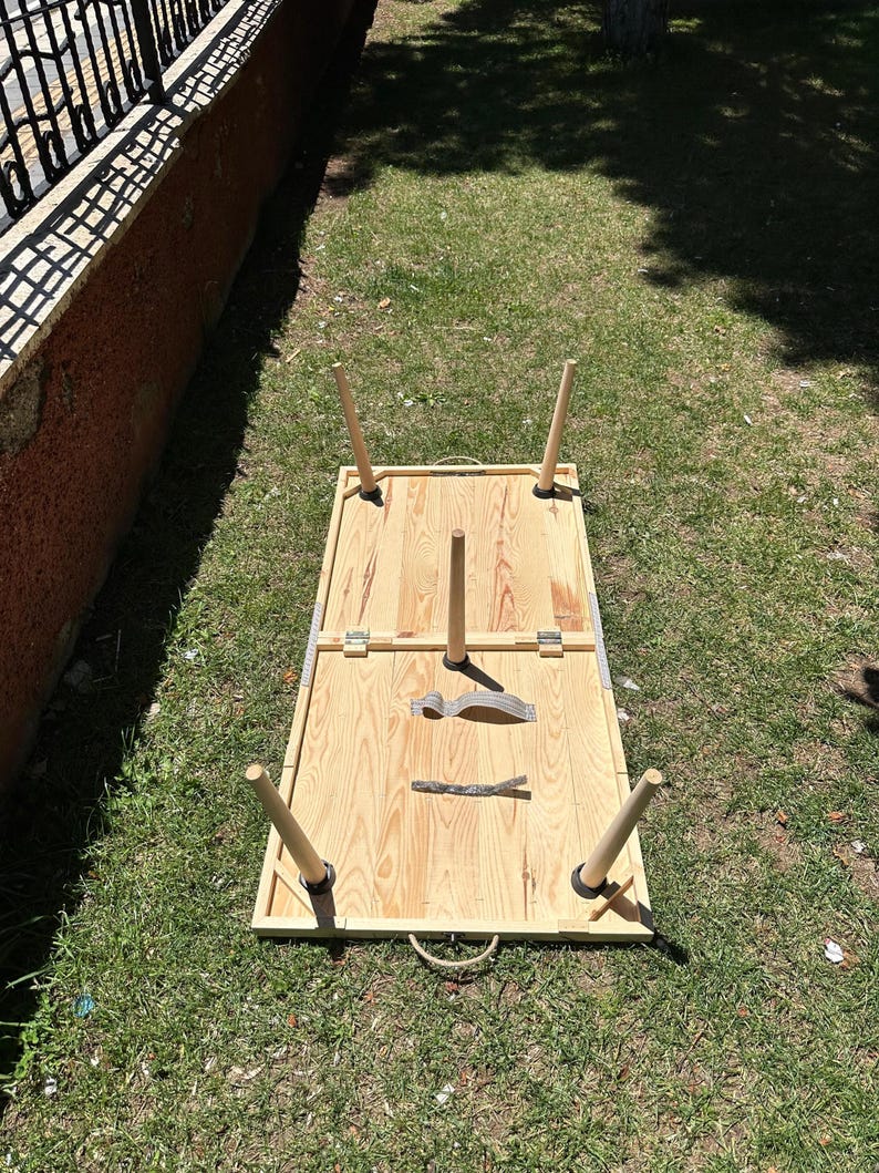May include: A rectangular wooden picnic table with four detachable legs. The table is made of light-colored wood and has a rope handle. A fork and knife are placed on the table. The table is set on a grassy area.