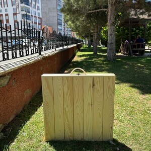 May include: A rectangular wooden folding table made of light-colored wood, with a woven handle. The table is standing upright on a grassy area, with a fence and buildings in the background.