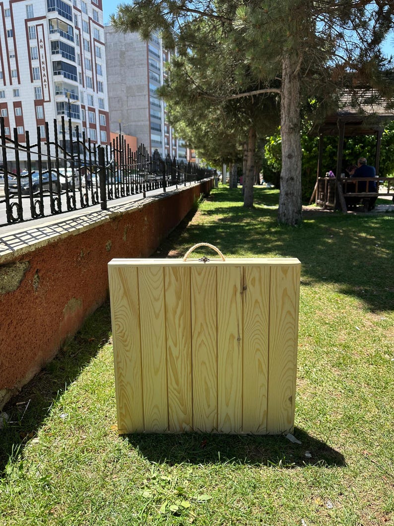 May include: A rectangular wooden folding table made of light-colored wood, with a woven handle. The table is standing upright on a grassy area, with a fence and buildings in the background.