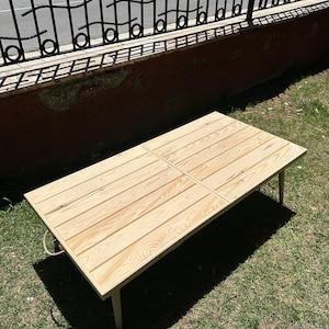 May include: A light-colored wooden table with a rectangular top and four legs. The tabletop is made of wooden planks arranged in a grid pattern. The table is outdoors on a grassy surface.
