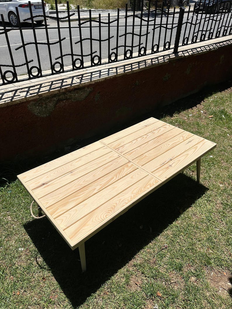 May include: A light-colored wooden table with a rectangular top and four legs. The tabletop is made of wooden planks arranged in a grid pattern. The table is outdoors on a grassy surface.