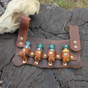 Könnte beinhalten: Ein brauner Leder-Trankgürtel mit vier kleinen Glasflaschen, jede mit einem unterschiedlich farbigen Verschluss. Der Gürtel ist mit Messingnieten befestigt und hat einen Riemen zum Tragen. Der Text "Green's Handcrafting" ist sichtbar.