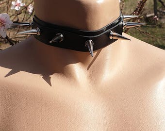 May include: Black leather choker necklace with multiple silver-coloured spikes. The choker is displayed on a mannequin, with a blurred background of flowering branches.