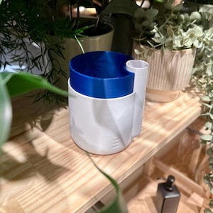 May include: A two-tone white and blue planter with a cylindrical shape. The top portion is a vibrant blue, while the base is white. A smaller, attached cylinder extends from the side. The planter is designed to hold plants and is placed on a wooden shelf.