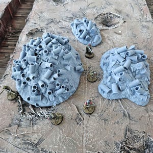 May include: Three gray, irregular-shaped terrain pieces, each covered in numerous gray bullet casings. Miniature figurines are placed around the terrain. The background is a textured, light brown surface, suggesting a battlefield setting.