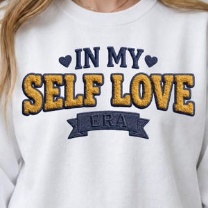Puede incluir: Sudadera blanca con la inscripción bordada "IN MY SELF LOVE ERA". Las letras son azul marino y doradas, con un efecto texturizado y difuso. Corazones azul marino flanquean "IN MY".