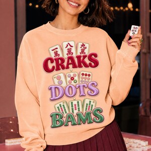 Peut inclure: Un sweat-shirt à col rond couleur pêche arborant un motif de tuiles de mah-jong et du texte. Le design comprend trois rangées de tuiles avec les mots CRAKS en rouge, DOTS en violet et BAMS en vert, accompagnés d'illustrations de tuiles.