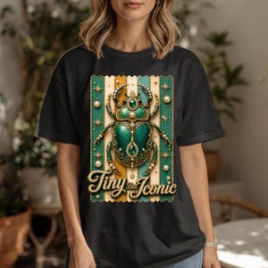 May include: A black t-shirt featuring a graphic of a green, jewel-encrusted beetle centered on a background of vertical gold and teal stripes. The beetle is adorned with pearls and small stars. Below the beetle, the text reads Tiny But Iconic.
