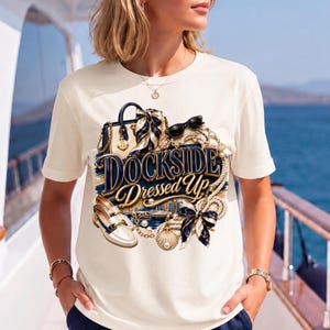 May include: A cream-colored short-sleeve t-shirt featuring a graphic print of a handbag, sunglasses, loafers, ropes, and bows with the text DOCKSIDE DRESSED UP in navy blue and gold lettering. The design includes nautical elements like anchors.