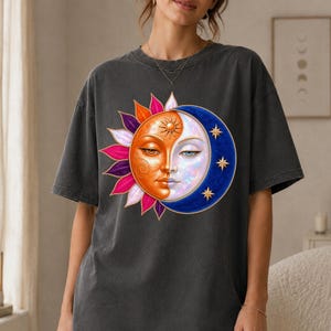 Peut inclure: Un t-shirt gris anthracite à manches courtes orné d'un grand motif représentant un visage de soleil et de lune. Le côté soleil est orange avec des rayons roses et violets, tandis que le côté lune est bleu avec des étoiles dorées.