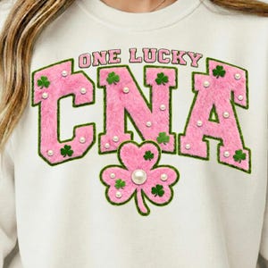 Peut inclure: Sweat-shirt blanc avec l'inscription "ONE LUCKY CNA" en rose, lettres en fausse fourrure soulignées de vert. Le motif comprend des trèfles et des perles, avec un grand trèfle en bas.