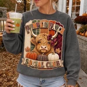 Peut inclure: Un sweat-shirt gris anthracite à col rond orné d'un motif de vache Highland avec un nœud bordeaux sur la tête. Le design comprend le texte PUMPKIN PATCH SOCIAL CLUB, des motifs à carreaux et trois citrouilles orange, à carreaux et crème.