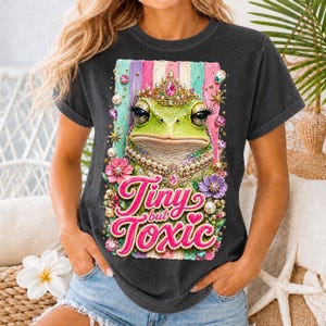 May include: Dark gray t-shirt featuring a cartoon frog wearing a crown and pearls, with the text "Tiny but Toxic" in pink and white. The frog is surrounded by flowers and jewels. The shirt has rolled-up sleeves.
