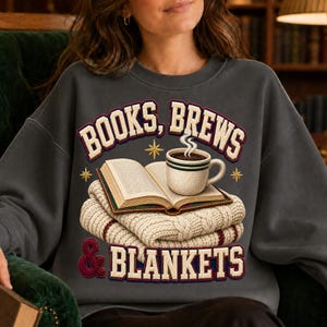 Peut inclure: Un sweat-shirt à col rond gris anthracite avec une impression graphique représentant un livre ouvert posé sur une couverture en tricot torsadé crème pliée et une tasse de café fumante. Le texte indique BOOKS, BREWS & BLANKETS.