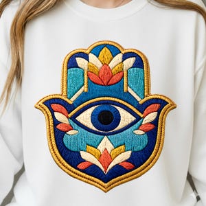 Puede incluir: Sudadera blanca con un diseño bordado de la mano de Fátima. La mano de Fátima es en tonos azules, amarillos, naranjas y blancos, con un motivo de ojo central. El diseño está delineado con hilo dorado.