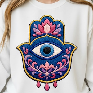 Puede incluir: Sudadera blanca con un diseño bordado de la mano de Hamsa. La Hamsa es azul con un ojo central, rodeado de detalles florales rosas, morados y dorados. El diseño incluye tres lágrimas rosas en la parte inferior.