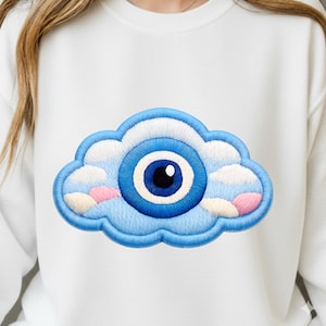 May include: White sweatshirt with an embroidered cloud design and a blue evil eye. The cloud has a blue outline, white and pink accents, and a central blue eye with a black pupil. A casual, everyday garment.