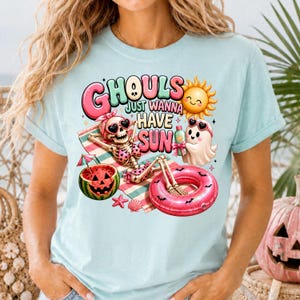 Peut inclure: T-shirt bleu clair avec l'inscription "Ghouls Just Wanna Have Sun". Le motif représente un squelette, un fantôme et un soleil profitant d'une journée à la plage avec une pastèque, une bouée et une boisson. Un design amusant sur le thème de l'été.