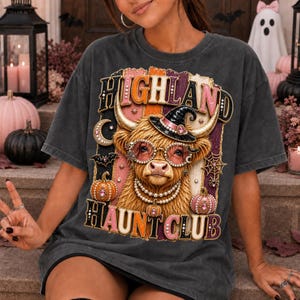 Peut inclure: Un t-shirt gris anthracite orné d'un motif de vache Highland portant un chapeau de sorcière, un collier de perles et des lunettes à bijoux. Le design inclut des citrouilles, une toile d'araignée et une chauve-souris, avec le texte HIGHLAND HAUNT CLUB.