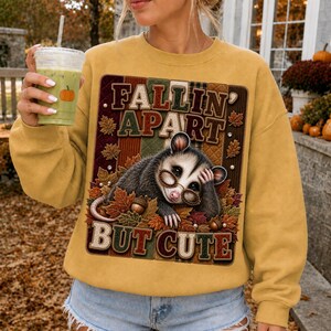 Könnte beinhalten: Ein senfgelbes Langarm-Sweatshirt mit einer Grafik eines Opossums mit Brille, umgeben von Herbstblättern und Eicheln. Der Text "FALLIN APART BUT CUTE" ist in Blockschrift über und unter der Opossum-Illustration aufgedruckt.