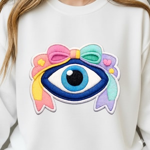 Puede incluir: Parche bordado con un gran ojo azul con iris azul claro y pupila negra. El ojo está rodeado por un diseño colorido con un lazo rosa, detalles amarillos y verdes y cintas de colores pastel. El parche está sobre un fondo blanco.