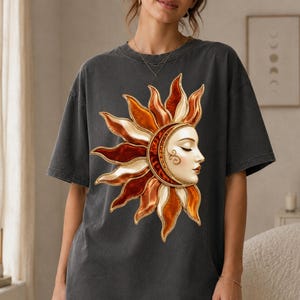 Peut inclure: Un t-shirt gris anthracite à manches courtes arborant un grand motif graphique représentant un soleil stylisé avec un visage humain. Le design du soleil intègre des tons chauds orange, dorés et crème, avec des détails tourbillonnants.