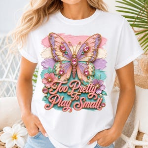 Peut inclure: Un t-shirt blanc avec une impression graphique représentant un papillon orné de bijoux roses et violets. Le motif comprend des coups de pinceau pastel, des accents floraux et le texte Too Pretty To Play Small en lettres cursives roses.