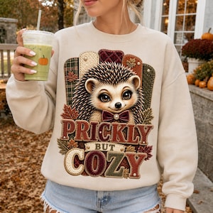 Peut inclure: Un sweat-shirt beige à col rond orné d'un hérisson portant un nœud papillon, entouré de panneaux style matelassé aux couleurs automnales. Le texte PRICKLY BUT COZY est écrit en lettres texturées, agrémenté de petites feuilles d'automne.