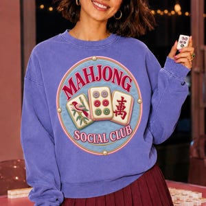 Peut inclure: Un sweat-shirt à col rond violet avec un motif circulaire portant l'inscription "MAHJONG SOCIAL CLUB" en rose. Le design présente trois tuiles de mah-jong, dont une avec un oiseau, une avec six points et une avec un caractère chinois rouge.