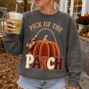 Peut inclure: Un sweat-shirt gris anthracite à col rond orné d'un motif de citrouille orange matelassée avec une anse en chaîne dorée et un nœud à carreaux. Le texte PICK OF THE PATCH est inscrit en lettres dorées et crème au centre du vêtement.