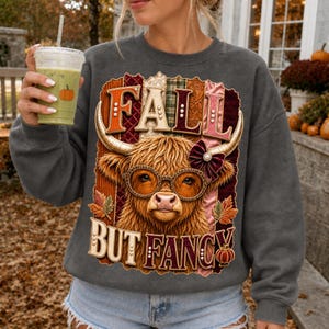 May include: A charcoal grey long-sleeve sweatshirt featuring a graphic of a Highland cow wearing leopard-print glasses and a maroon bow. The text reads FALL BUT FANCY, surrounded by vertical textured panels in orange, brown, and pink tones.