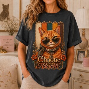 Peut inclure: Un t-shirt gris anthracite orné d'un motif représentant un chat tigré orange portant des lunettes de soleil et un foulard à motifs. Le chat est encadré par des bandes verticales et des fleurs, avec le texte "Chaotic But Adorable" en dessous.