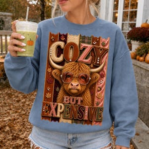 May include: A dusty blue crewneck sweatshirt featuring a graphic of a Highland cow wearing glasses. The text reads COZY BUT EXPENSIVE. The design is set against a background of vertical plaid panels with autumn leaves and small pearl accents.