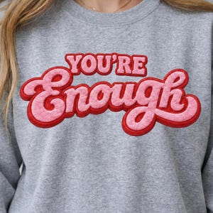 Peut inclure: Un sweat-shirt gris avec l'inscription "YOU'RE Enough" dans une police rétro et arrondie. Le texte est rose avec un contour rouge, créant un design audacieux. Le sweat-shirt est de style décontracté et confortable.