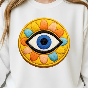 May include: White sweatshirt featuring a large, embroidered eye design. The eye is blue with a black outline, surrounded by a yellow circle with colorful petal-like shapes in orange, yellow, and blue. The design is centered on the chest.