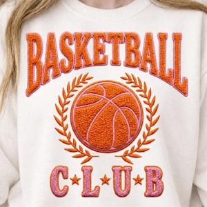 Puede incluir: Sudadera de color crema con las palabras "BASKETBALL CLUB" en letras bordadas en naranja y rosa. Una pelota de baloncesto está centrada dentro de una corona de laurel. La sudadera es una prenda deportiva informal.