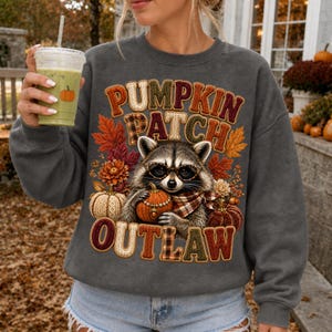 Peut inclure: Un sweat-shirt gris anthracite à col rond orné d'un motif de raton laveur portant des lunettes et une écharpe à carreaux, tenant une citrouille. Le texte PUMPKIN PATCH OUTLAW est imprimé dans une police texturée et colorée avec des feuilles d'automne.